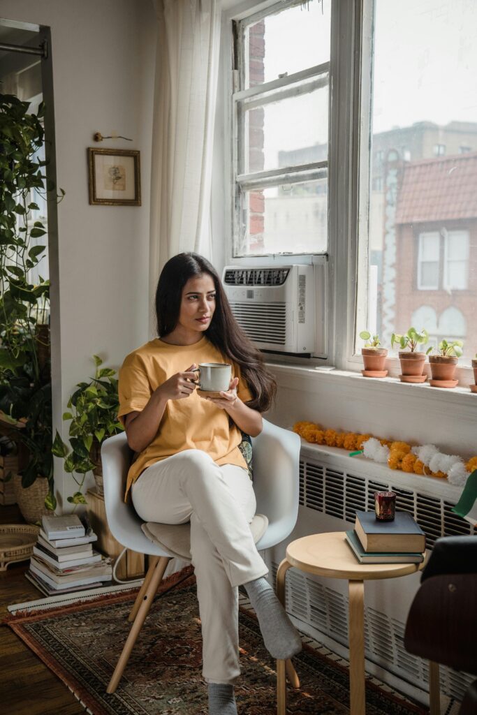 Cozy bedroom reading corner with woman sitting by the window holding a cup of tea, surrounded by books and plants