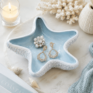 Blue ceramic starfish jewelry dish holding rings and earrings on a coastal vanity with seashells, candle, and beach-inspired home decor.