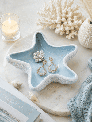 Blue ceramic starfish jewelry dish holding rings and earrings on a coastal vanity with seashells, candle, and beach-inspired home decor.