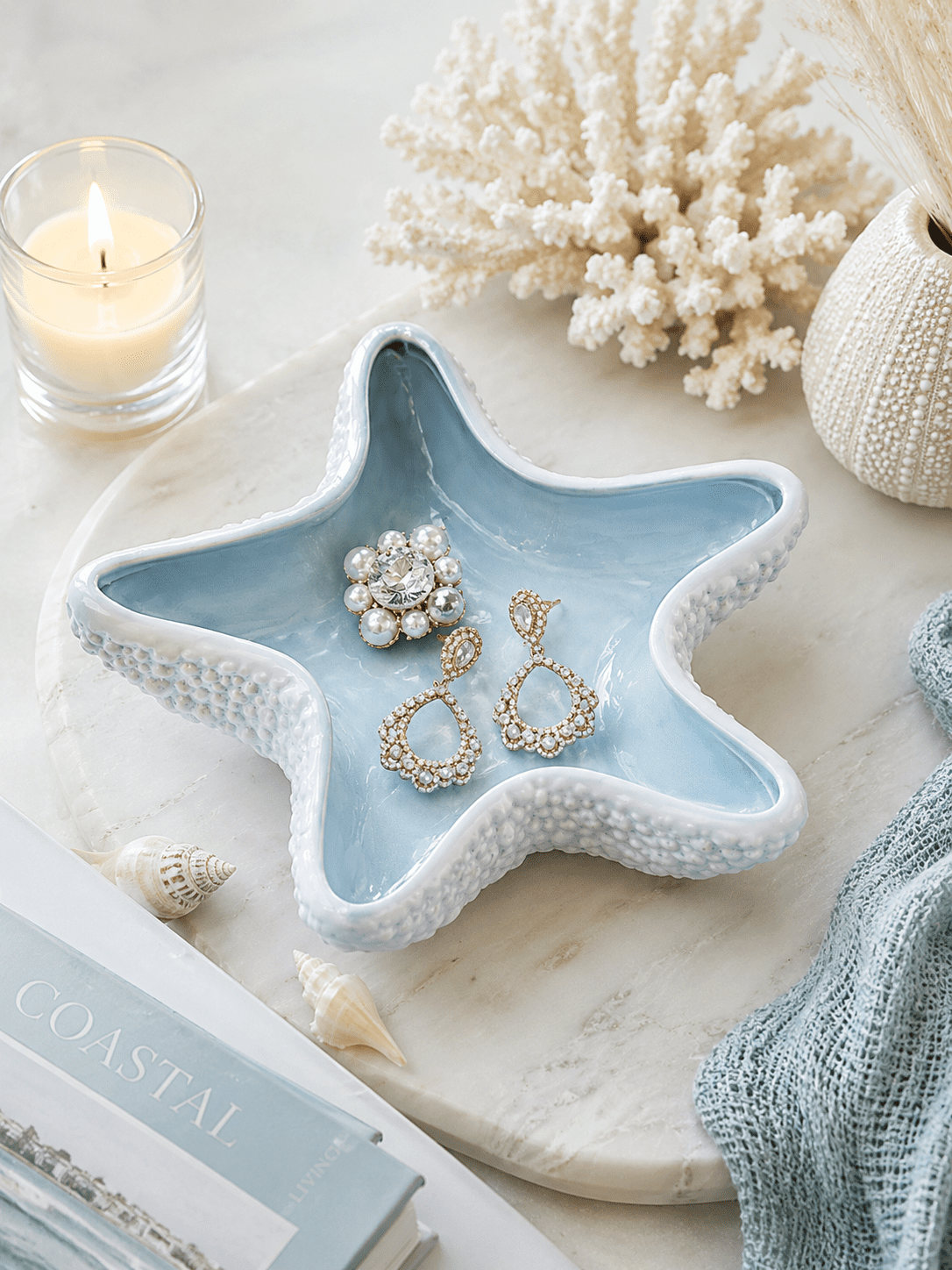 Blue ceramic starfish jewelry dish holding rings and earrings on a coastal vanity with seashells, candle, and beach-inspired home decor.