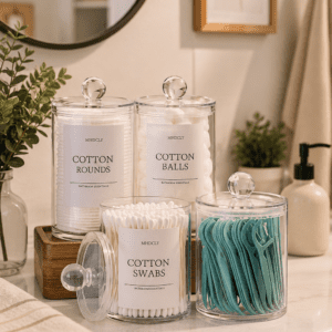 Clear acrylic bathroom storage canister set with cotton rounds, cotton balls, cotton swabs, and dental floss picks displayed on a modern bathroom vanity.