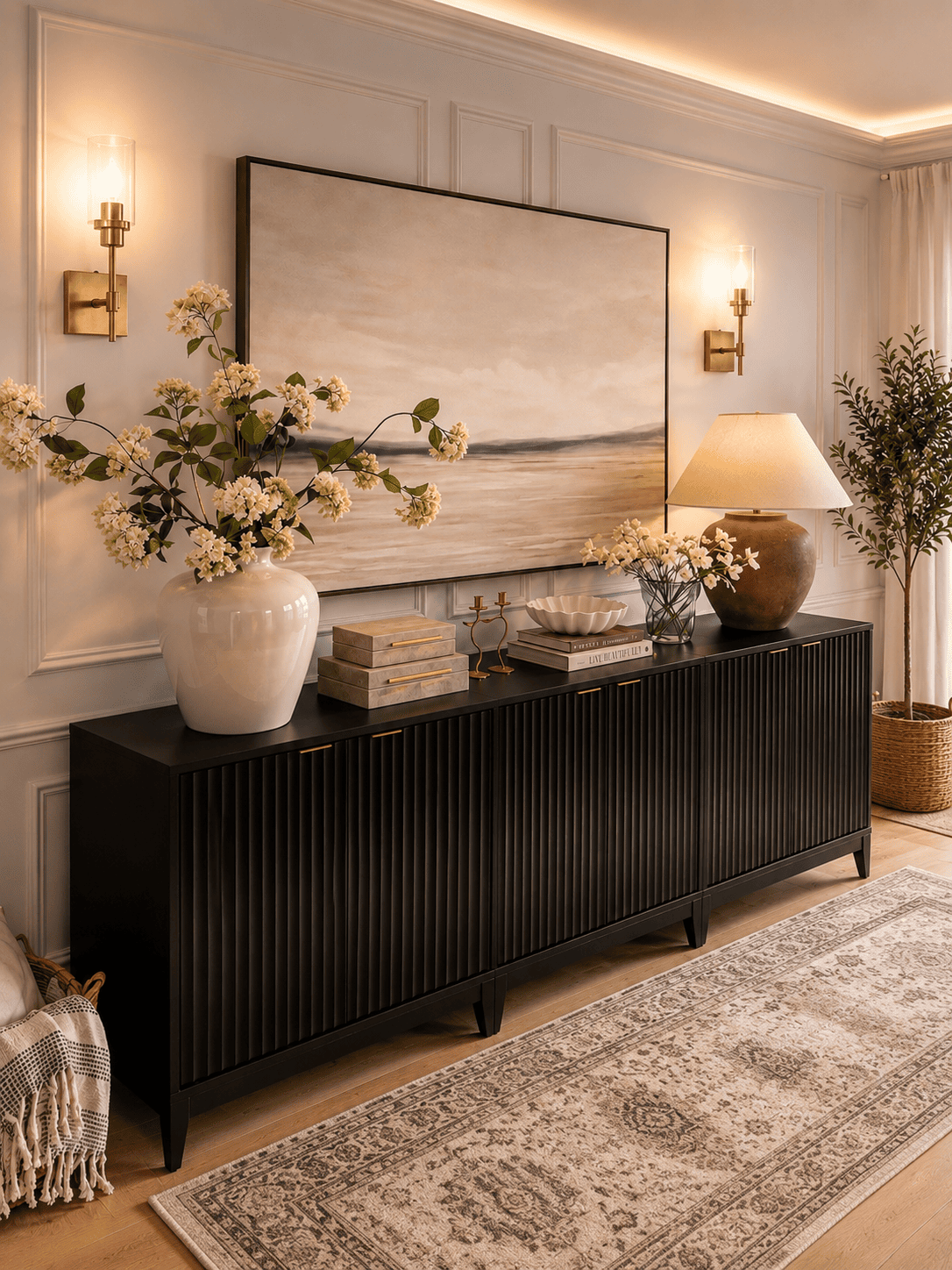 Modern living room featuring a black fluted sideboard cabinet, oversized wall art, table lamp, wall sconces, ceramic vases, faux olive tree, decorative books, candles, and neutral home décor.