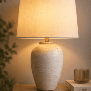 Neutral ceramic bedside table lamp with a linen shade casting warm ambient lighting on a wooden nightstand in a cozy bedroom.