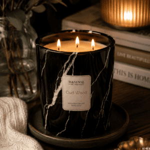 Luxury black marble triple wick Oud Wood scented candle on a decorative tray with cozy knit blanket, books, and elegant home decor in a warm living room.