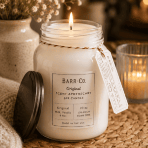 Large white apothecary scented jar candle with a rustic lid, surrounded by cozy blankets, dried flowers, and soft candlelight in a warm farmhouse-style living room.