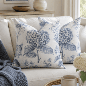 Blue botanical floral throw pillow covers on a neutral linen sofa with coastal-inspired decor, woven tray, soft knit blanket, and natural wood accents in a bright farmhouse living room.