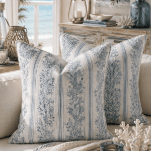 Blue coastal floral throw pillow covers styled on a neutral sofa with beach-inspired decor, candles, woven textures, and an ocean view creating a relaxing coastal living room.