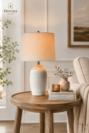 Neutral ceramic table lamp with beige linen shade styled on a round wood side table with books, greenery, candle, and cozy living room decor.