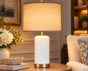 White ceramic table lamp with linen drum shade and gold-tone base styled on a round wood table in a cozy living room.