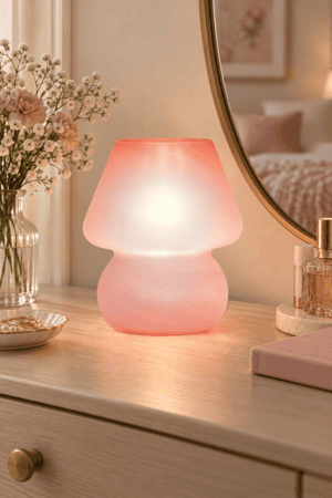 Blush pink glass table lamp glowing warmly on a light wood bedroom dresser beside flowers, perfume, jewelry, and round mirror.