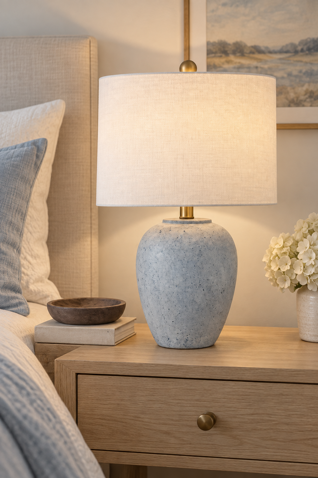 Light blue speckled ceramic table lamp with white linen drum shade and brass accents styled on a natural oak nightstand beside neutral bedding and white flowers.