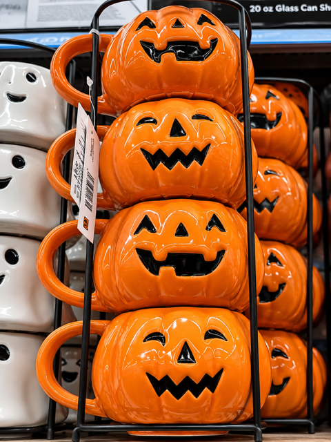 Glossy orange ceramic Jack-O’-Lantern pumpkin mugs with playful black Halloween faces displayed together in a black metal mug storage rack.