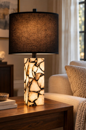 Black drum shade table lamp with glowing cream and dark patterned base on wooden table beside a beige upholstered sofa.